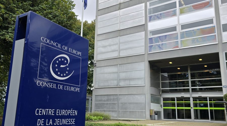 Photographie de l'extérieur du centre européen de la jeunesse à Strasbourg. Un panneau avec le logo du Conseil de l'Europe est présent au premier plan et annonce « Centre européen de la jeunesse / European Youth Center". À l’arrière-plan, l'entrée du centre est visible.